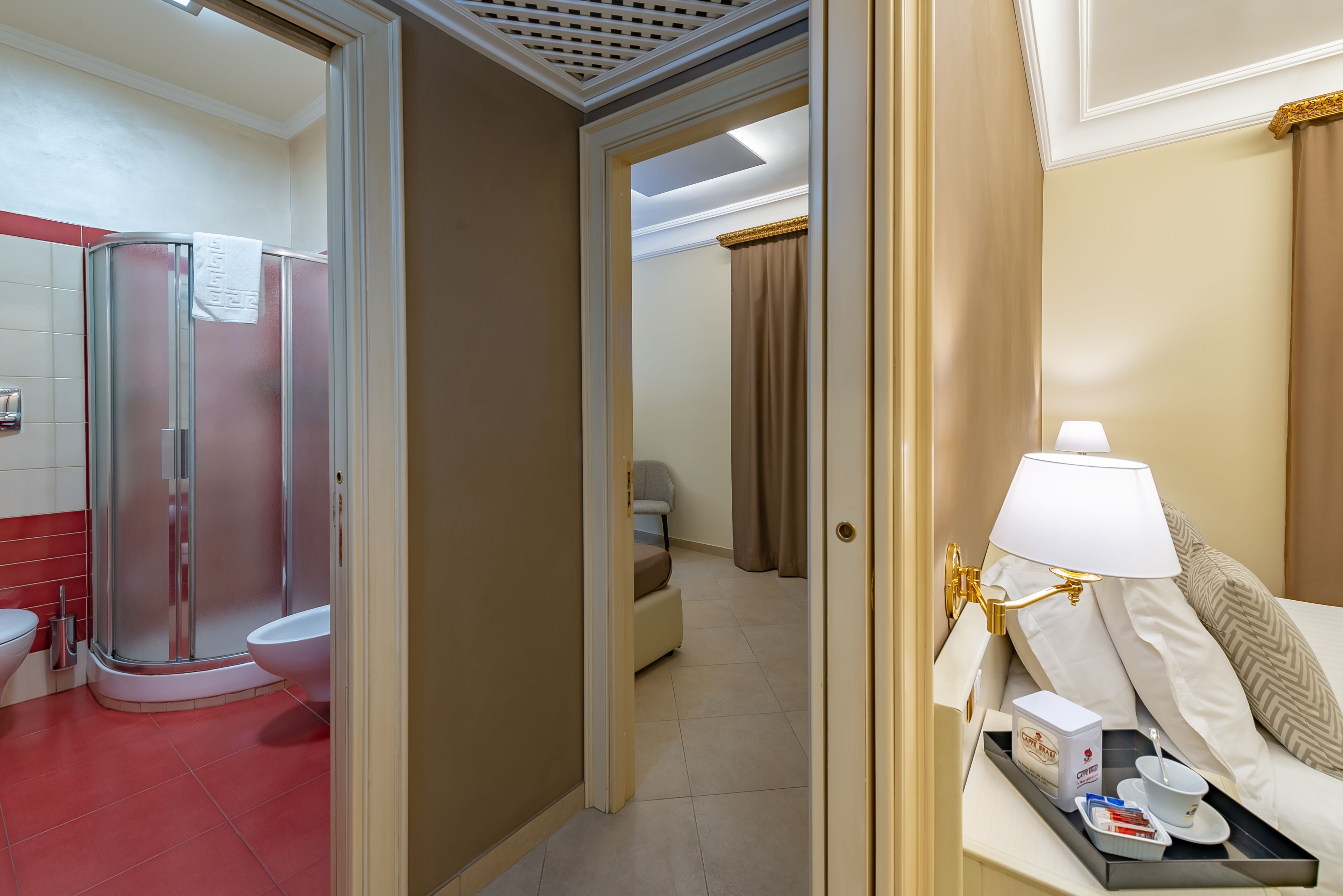 A hotel bathroom with a shower, toilet, and bidet. There is a bed with a bedside table, a lamp, and a tray with toiletries.
