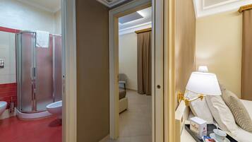 A hotel bathroom with a shower, toilet, and bidet. There is a bed with a bedside table, a lamp, and a tray with toiletries.