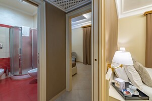A hotel bathroom with a shower, toilet, and bidet. There is a bed with a bedside table, a lamp, and a tray with toiletries.