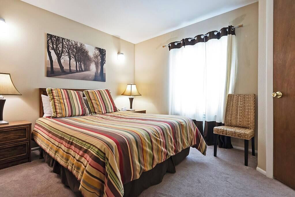Un dormitorio con cama, una silla, una lámpara, un cuadro y una ventana con cortinas.