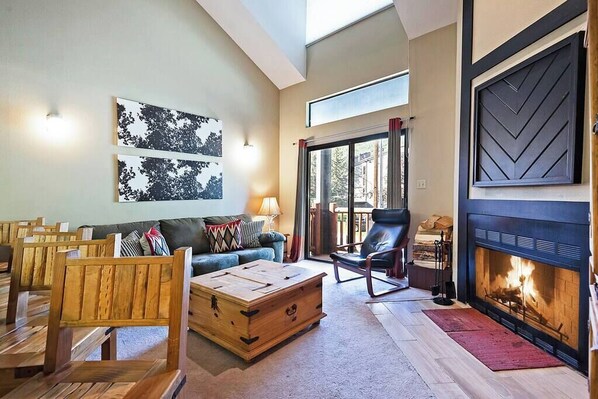 A living room with a fireplace, a sofa, a wooden coffee table, and a view through a glass door.