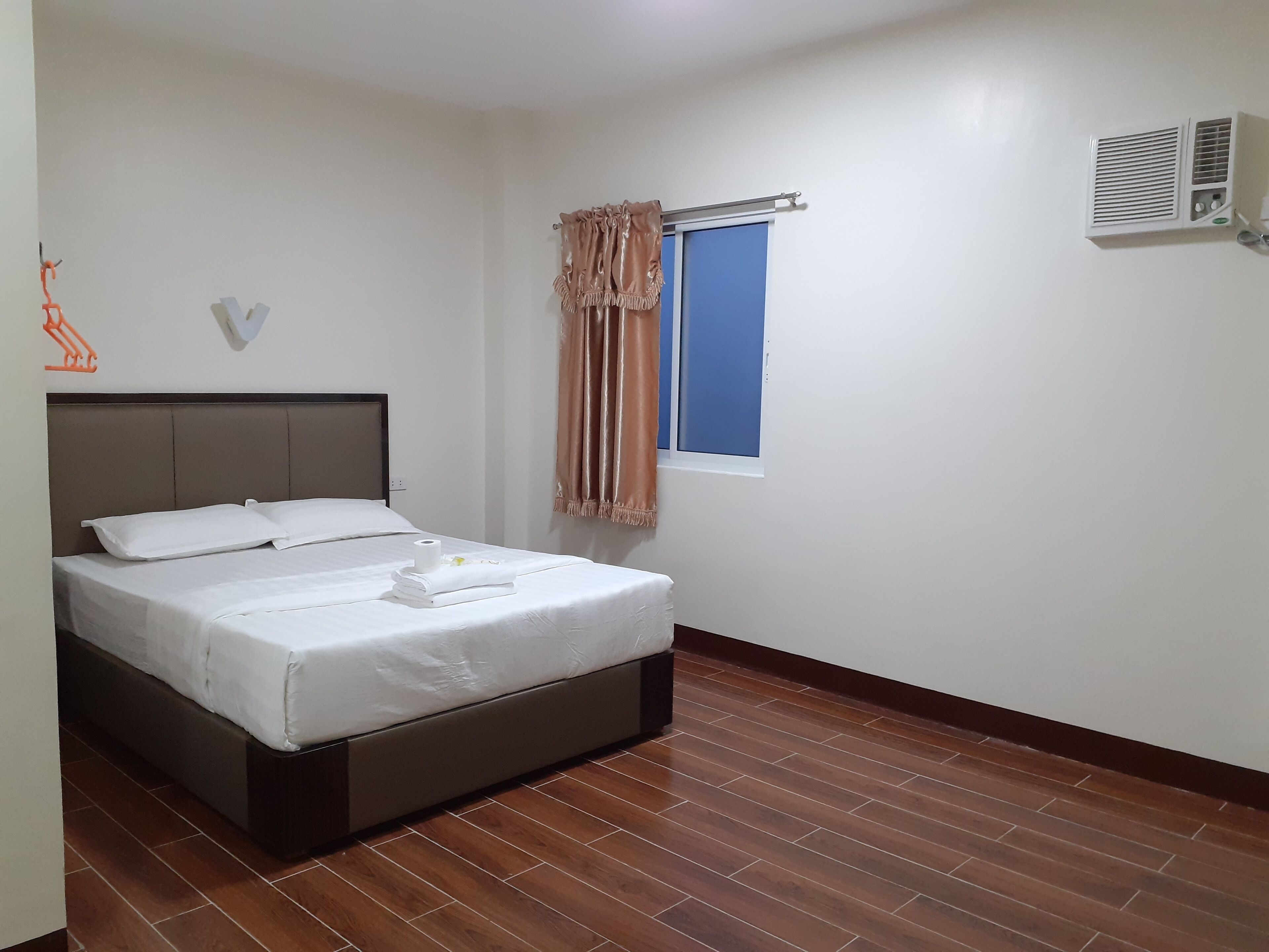 Una cama bien hecha con ropa de cama blanca y una toalla doblada, una ventana con cortinas y un equipo de aire acondicionado en la pared.