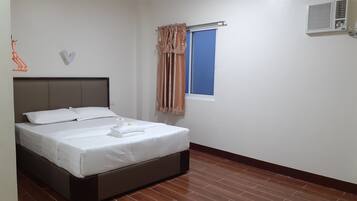 Una cama bien hecha con ropa de cama blanca y una toalla doblada, una ventana con cortinas y un equipo de aire acondicionado en la pared.