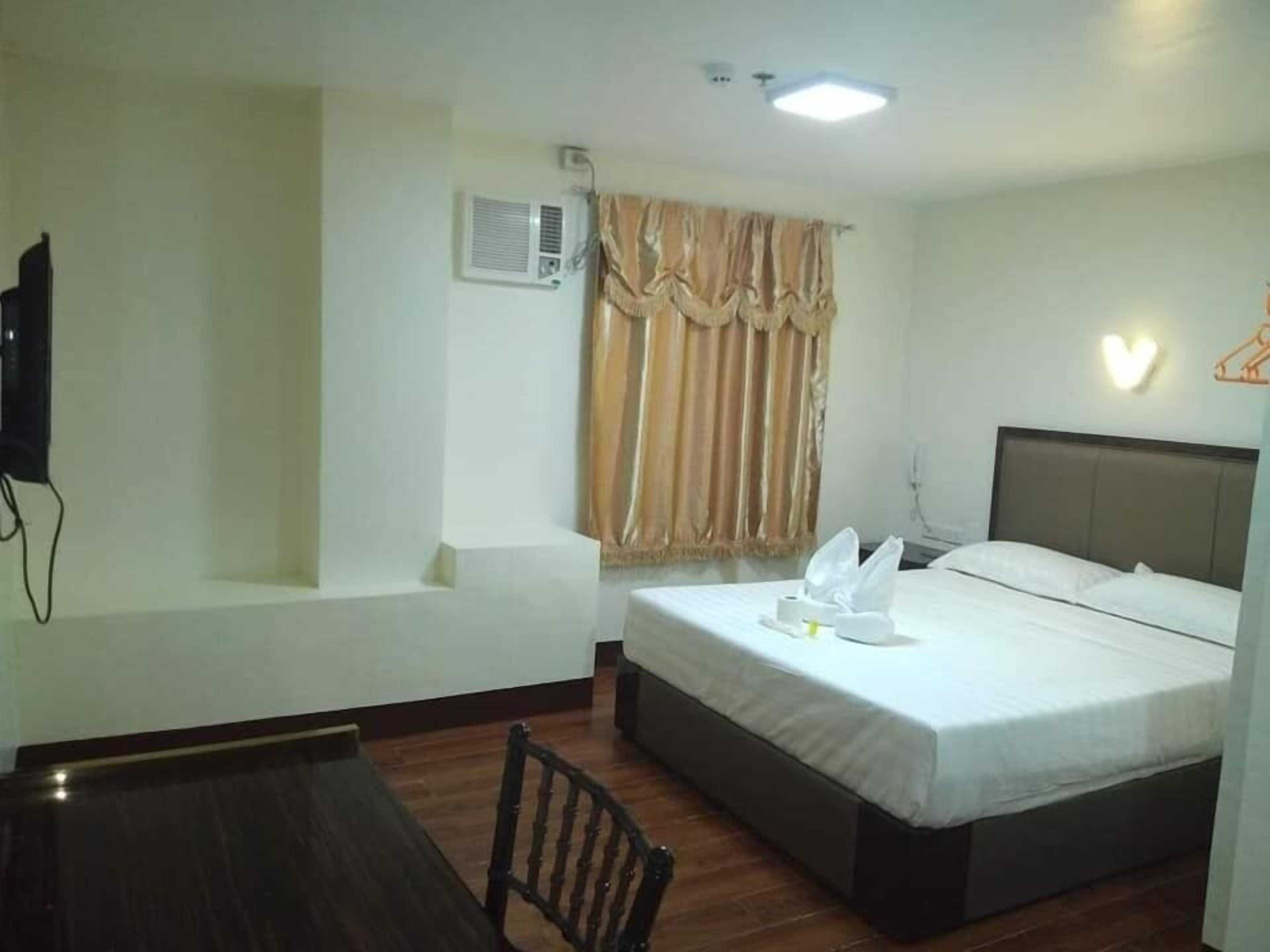 A hotel room with a bed, a desk, a chair, a TV, an air conditioning unit, and a window with curtains.