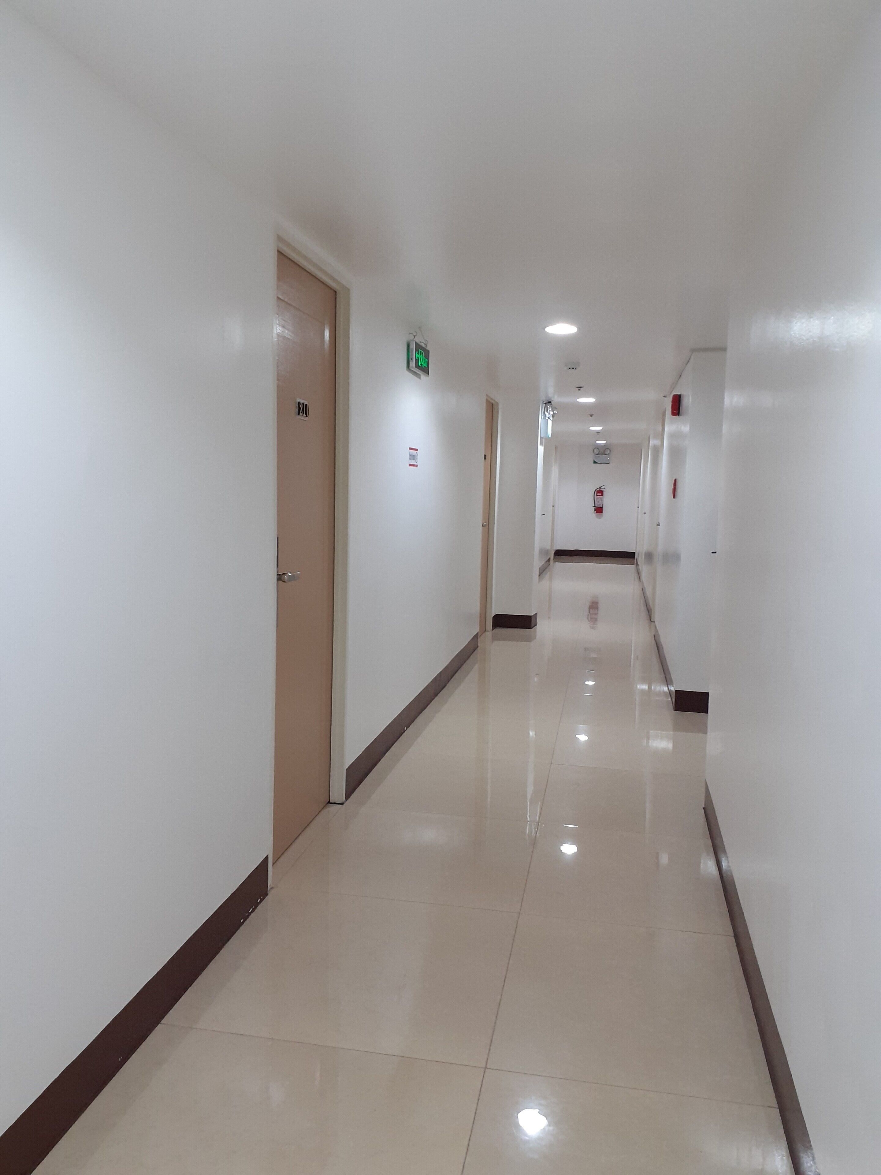 A clean, well-lit hotel corridor with white walls, a shiny tiled floor, and doors leading to rooms.