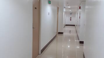 A clean, well-lit hotel corridor with white walls, a shiny tiled floor, and doors leading to rooms.
