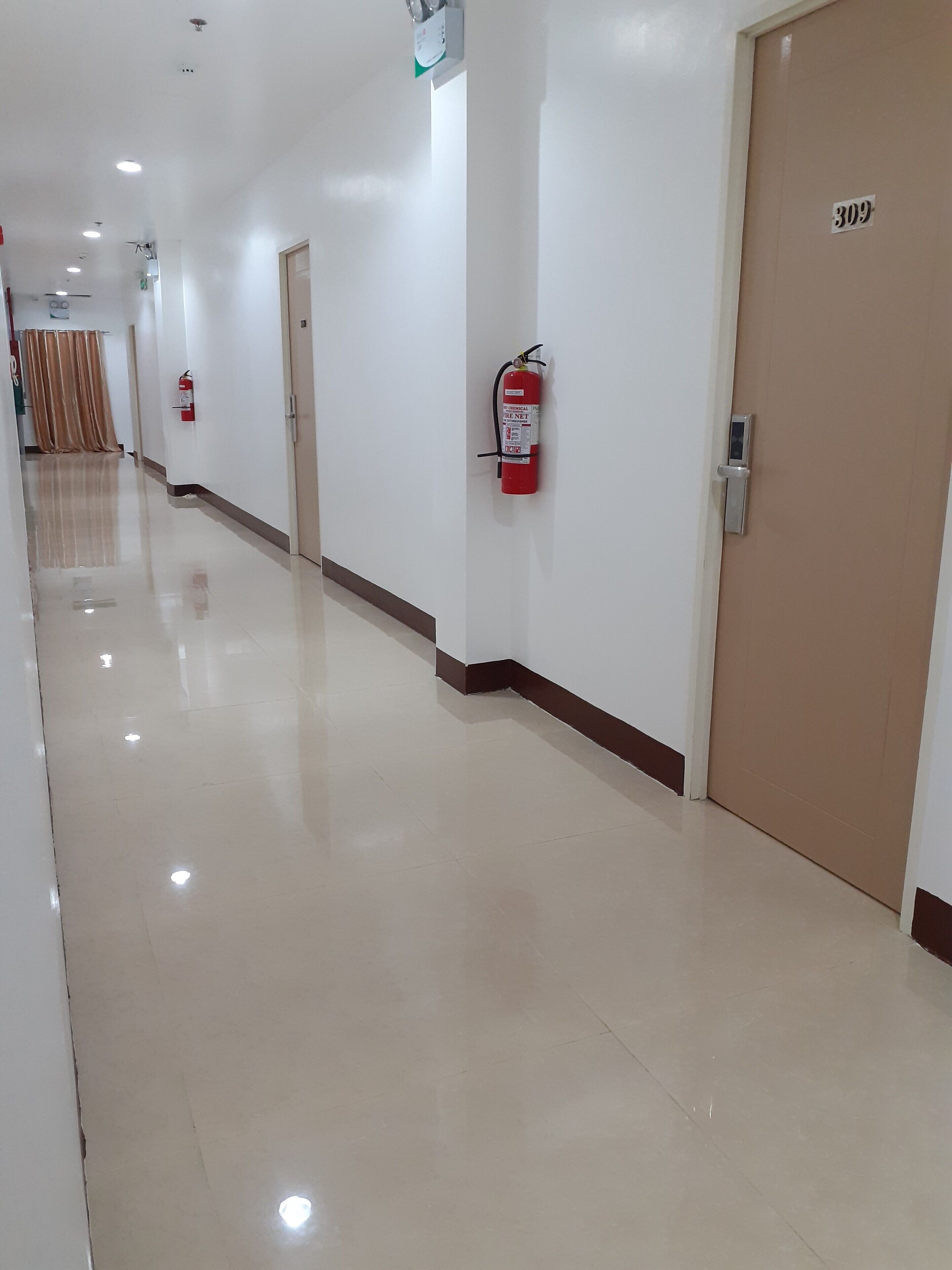 A hotel corridor with a fire extinguisher mounted on the wall, a door with a number sign, and a reflective floor.