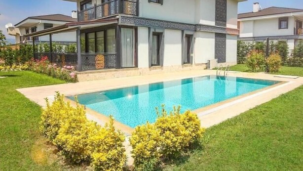 Una casa moderna de dos pisos con piscina, rodeada de césped y arbustos cuidados.
