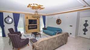 Una sala de estar con un sofá beige, un sofá azul, un sillón de cuadros, una mesita de centro, un televisor, una araña y un reloj en la pared.