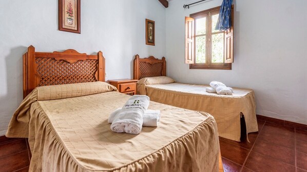 A hotel room with two beds, each with a wooden headboard and a beige bedspread. There are two nightstands, one between each bed, and a window with a wooden frame and a curtain. - Balcón del Río Sayalonga by Ruralidays (Málaga)