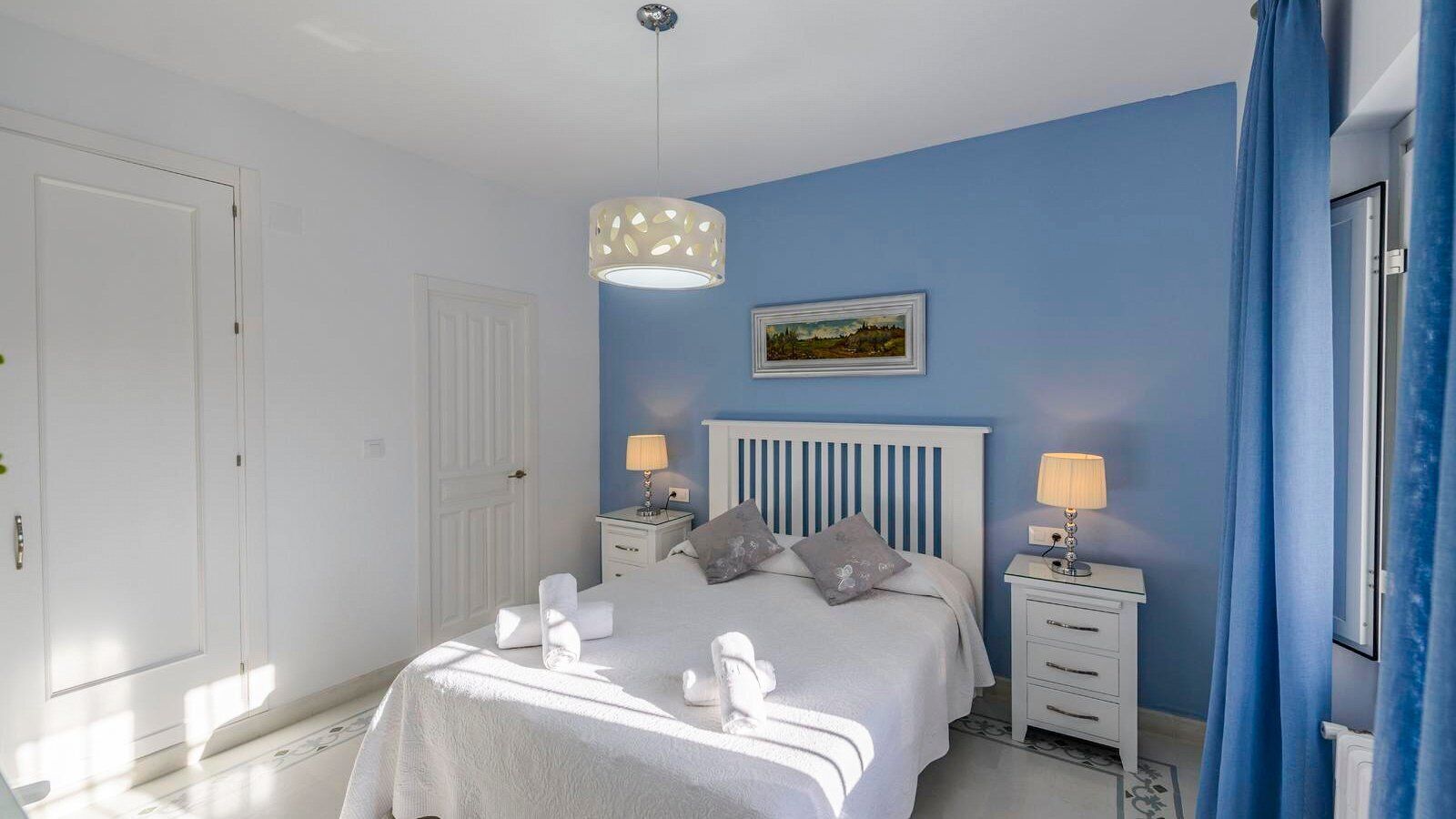 Un dormitorio con cama, dos mesitas de noche, una lámpara colgante, un cuadro en la pared y una puerta.