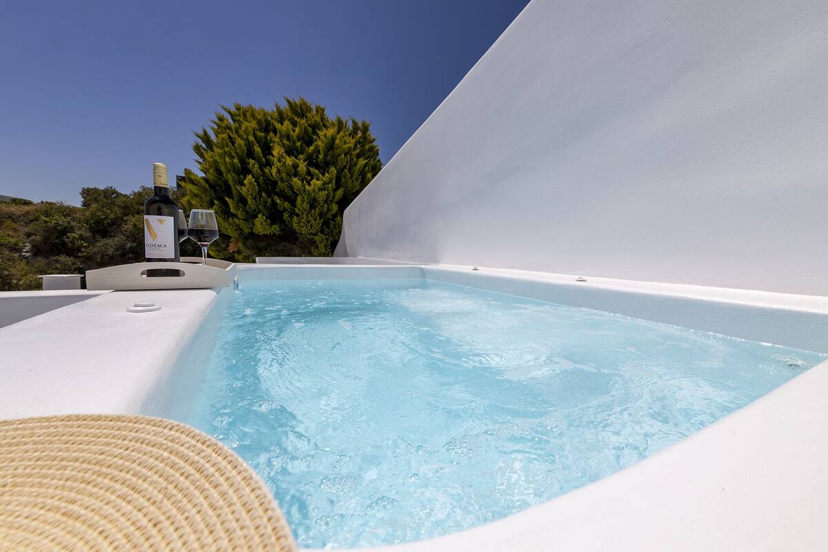 A hot tub with a bottle of wine and a glass, surrounded by white walls and a tree.