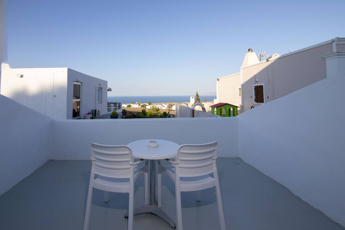 A white balcony with two chairs and a table, overlooking a coastal town with white buildings and a clear sky.