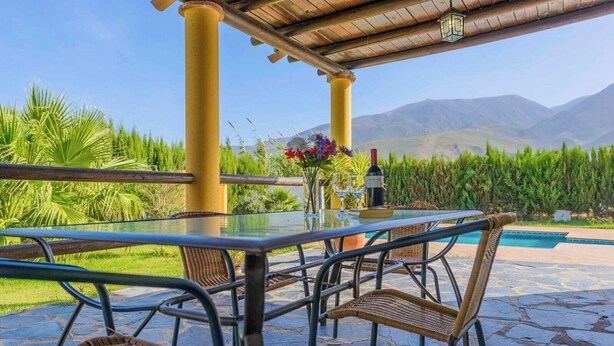 An outdoor dining area with a glass table, metal chairs, a bottle of wine, and a view of mountains. - La Chumbera Órgiva by Ruralidays (Granada)