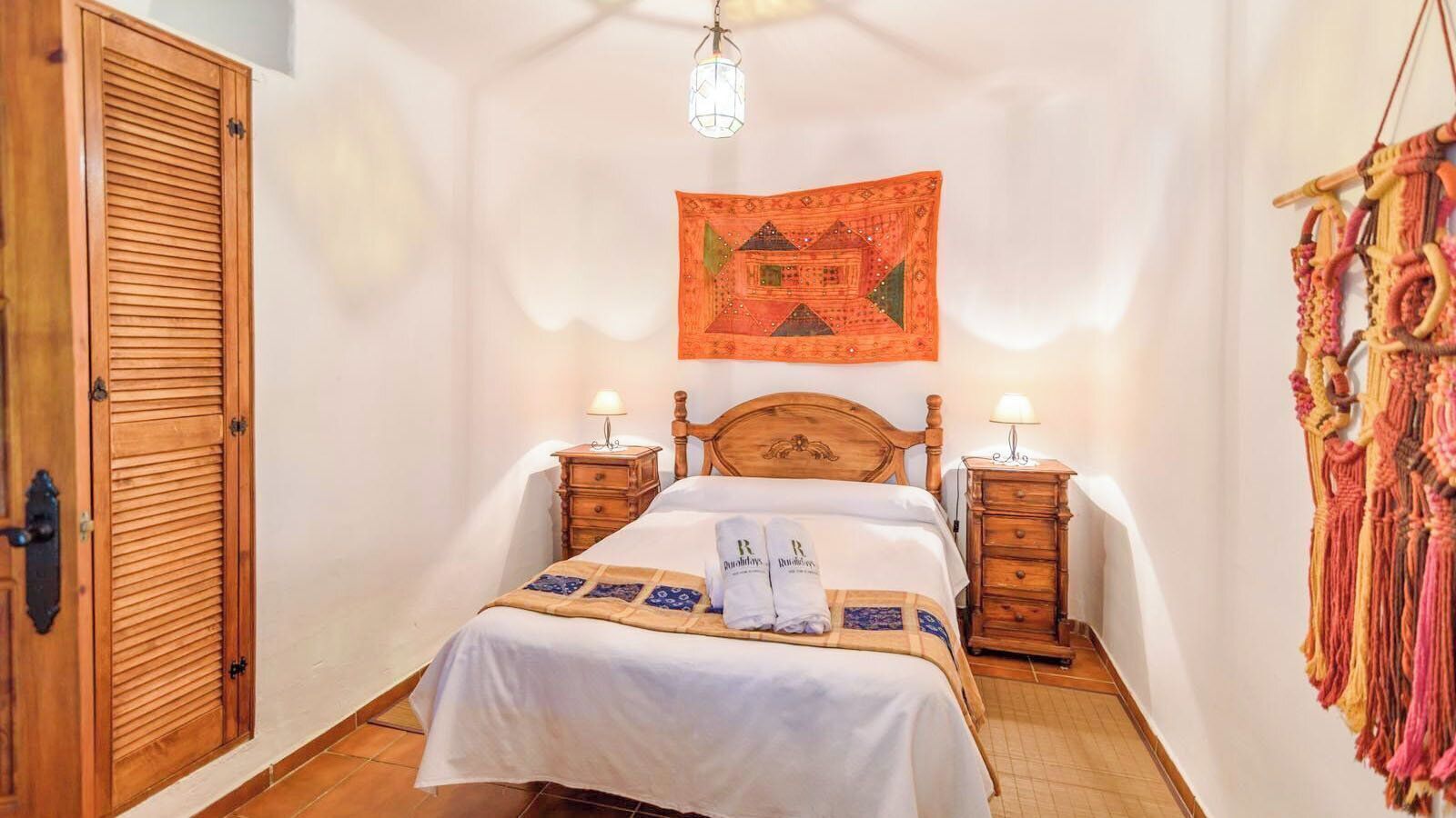 A bedroom with a wooden bed, two bedside tables with lamps, a wooden wardrobe, and a tapestry on the wall.