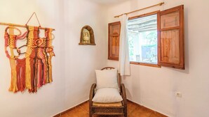 A room with a wicker chair, a macrame wall hanging, a small wooden shelf with a decorative item, and a window with wooden shutters.