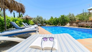 A poolside area with lounge chairs, a thatched umbrella, and an open book with sunglasses on a striped towel. - El Torcal Antequera - La Higuera by Ruralidays (Málaga)
