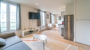 Un soggiorno moderno con un divano grigio, una TV a schermo piatto fissata al muro, un'area pranzo con un tavolo e sedie bianche e una cucina con frigorifero in acciaio inossidabile.