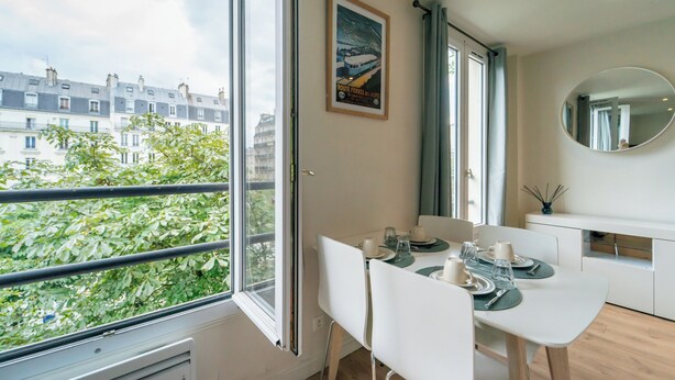 Un'area pranzo moderna con un tavolo apparecchiato per due, una grande finestra vista edifici e un poster incorniciato al muro.