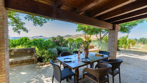 An outdoor patio with a table set for dining, surrounded by wicker chairs, a brick wall, and lush greenery.