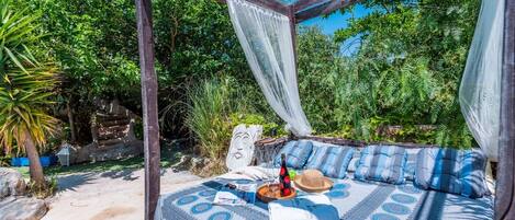 A covered outdoor seating area with a patterned tablecloth, cushions, and a bottle of wine.
