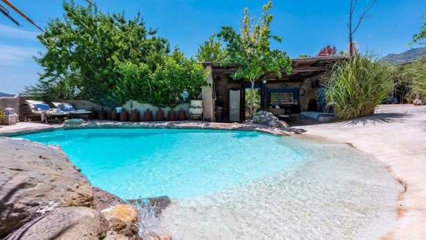 Piscina azul cristalina rodeada por pedra natural e vegetação, com pérgula de madeira e espreguiçadeiras.