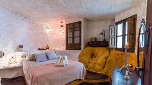 Um quarto rústico com uma cama, um banco de madeira, uma mesa com uma vela e uma janela com vista.