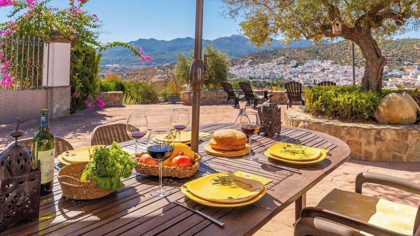 An outdoor dining area with a view of a hillside town, a table set with plates, glasses, and a basket of bread and vegetables.