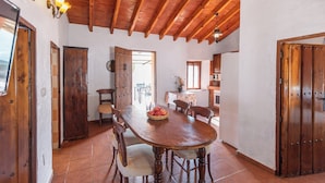 Zona de menjador amb taula de fusta, cadires i una cistada de fruita. Hi ha portes de fusta i un paviment de rajoles.