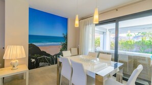 Un espace repas moderne avec vue sur la plage, un tableau représentant l’océan et un balcon donnant sur la verdure.