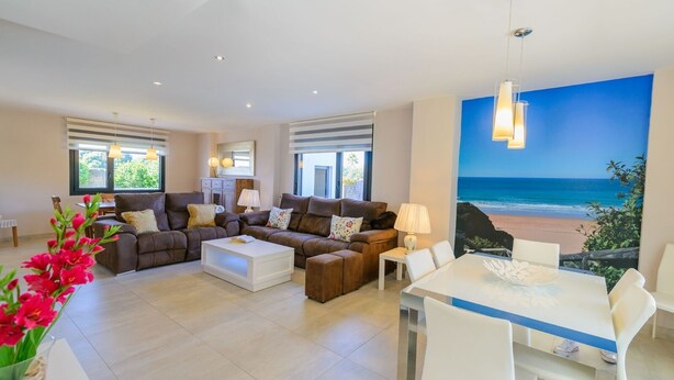 Un salon spacieux comprenant un grand canapé, une table basse et un coin repas avec une table et des chaises. Une grande fenêtre offre une vue sur la plage.