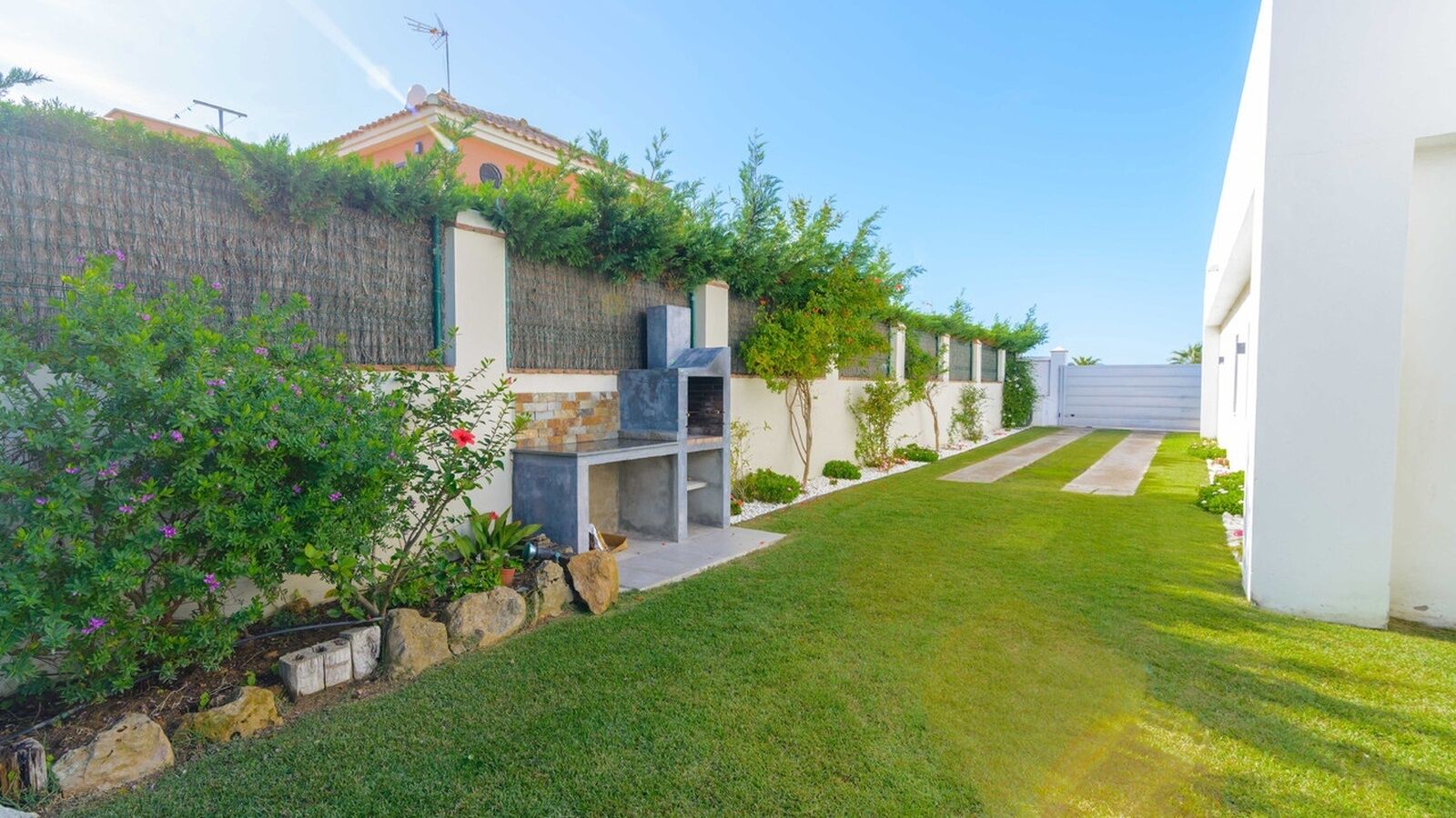A modern outdoor kitchen area with a stone wall, a grassy lawn, and a pathway leading to a white building.