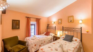 A room with two beds, a chair, a window with curtains, wall-mounted lamps, and framed pictures.
