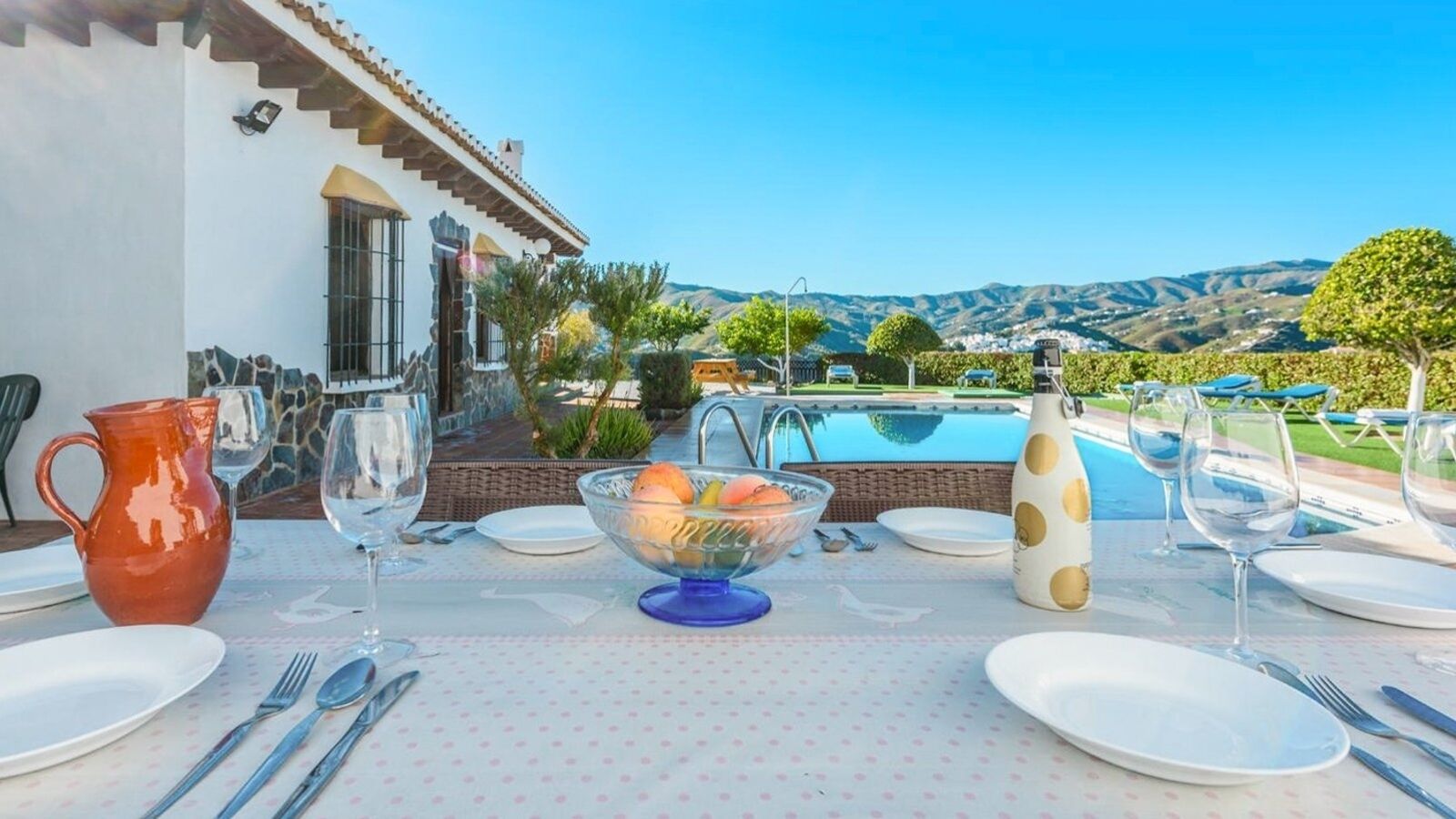 A dining table set outdoors with a pool and mountains in the background.