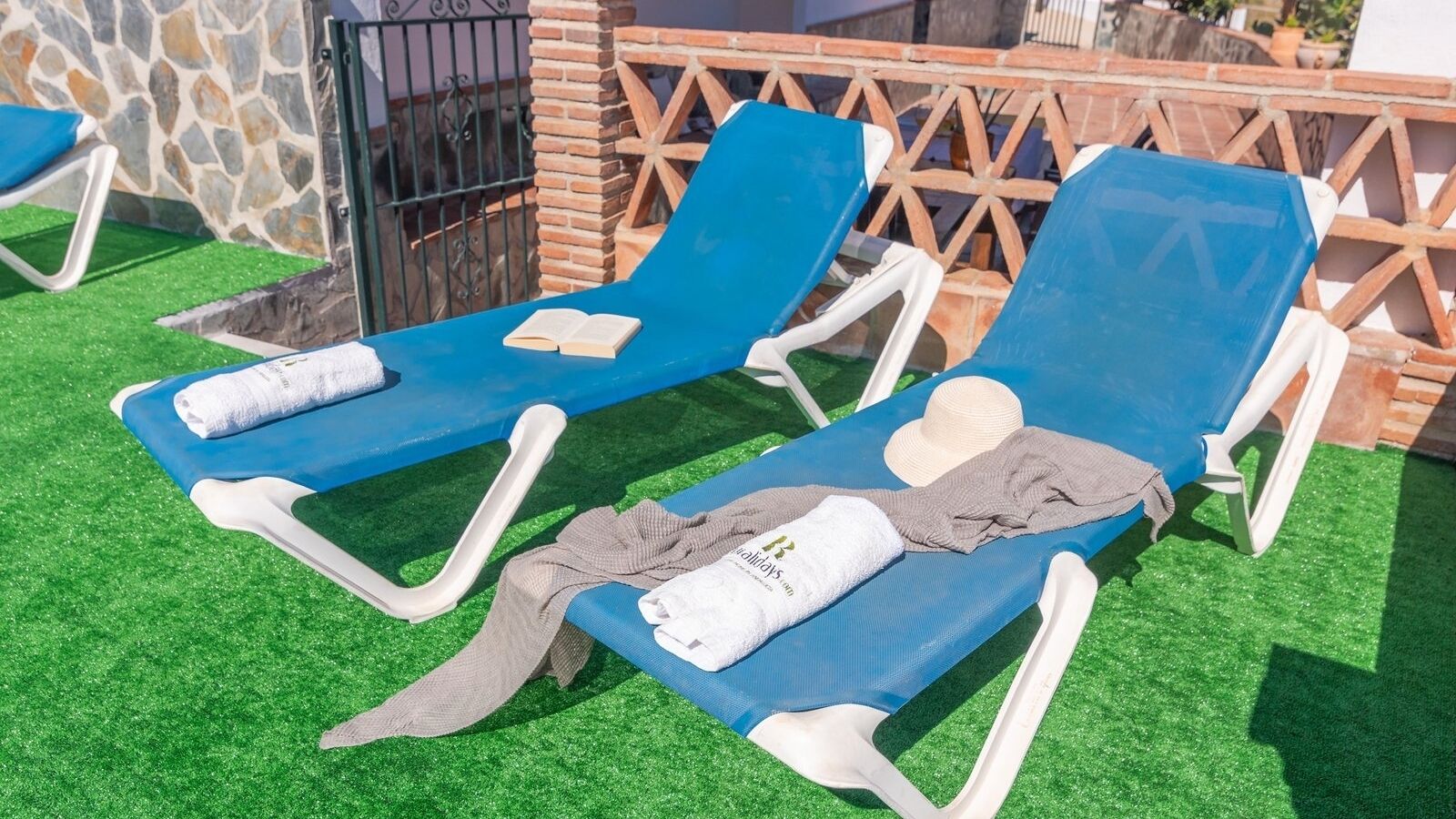 Deux fauteuils bleus, accompagnés de serviettes et d’un chapeau, sont placés sur un gazon artificiel.