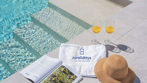 Une scène de bord de piscine avec une serviette, deux verres de jus d’orange, un chapeau de paille et une brochure.