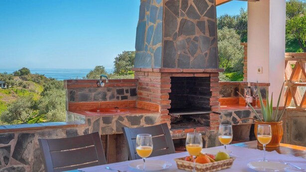 Un espace extérieur aménagé pour les repas, avec un barbecue en pierre, une table dressée avec des verres et un panier de fruits, et une vue sur la mer et les collines.
