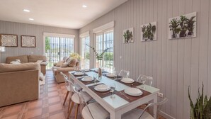 A dining area with a long table set for four, surrounded by chairs. There is a sofa and a window with a view of greenery outside.