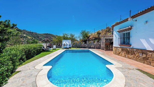 A rectangular swimming pool with stone edging, surrounded by a stone wall and a blue building with a tiled roof.