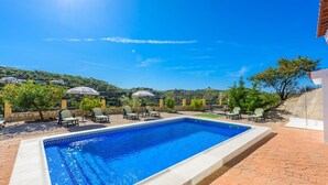 Une piscine rectangulaire bordée de carreaux, entourée de chaises longues et de parasols, avec en arrière-plan des collines et un ciel dégagé.