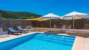 Une piscine entourée de chaises longues et de parasols, le tout bordé d’un mur élevé et avec une colline en arrière-plan.