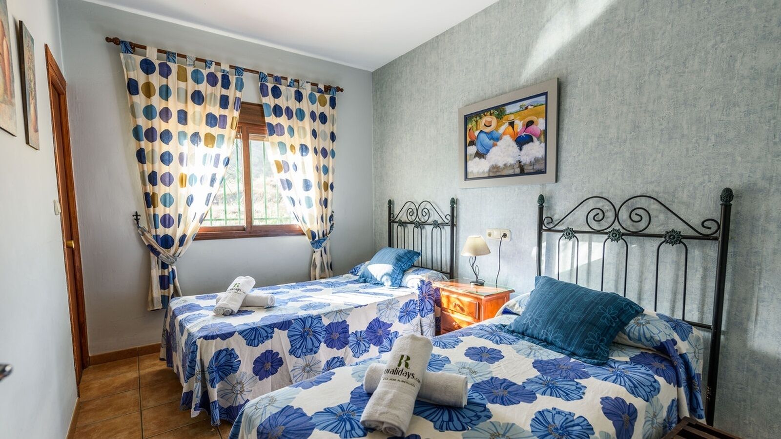 Une chambre avec deux lits, une fenêtre avec des rideaux, un tableau au mur et une table de chevet avec une lampe.