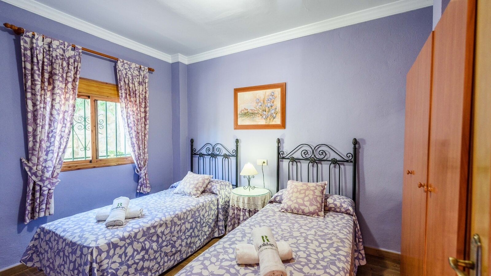 Un dormitorio con dos camas, una ventana con cortinas, una mesita de noche con lámpara y un cuadro en la pared.