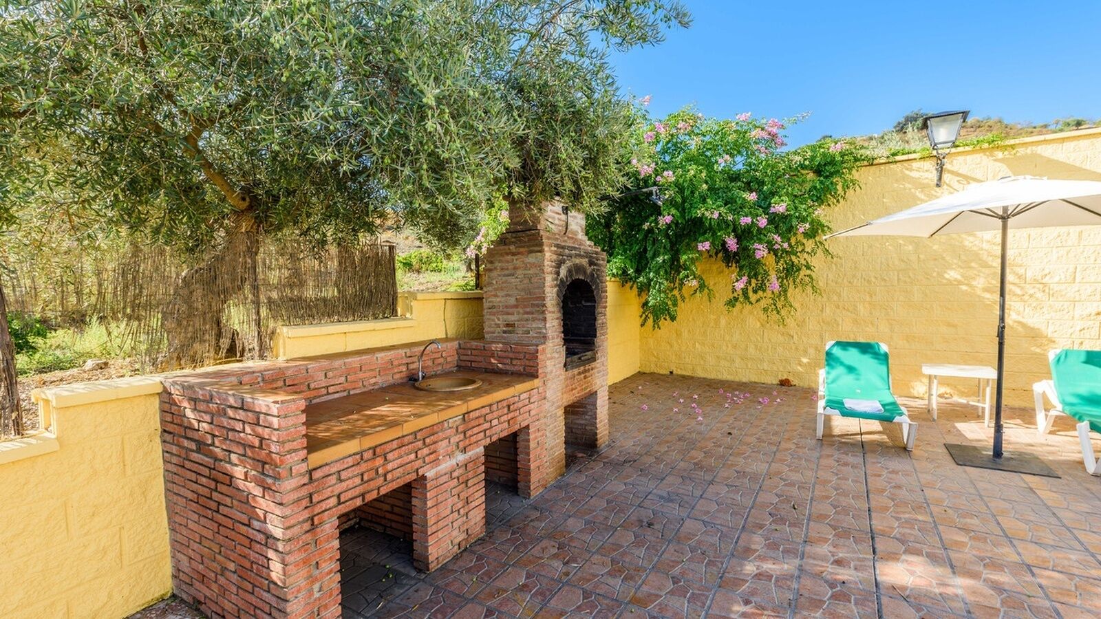 An outdoor patio with a brick barbecue grill, a yellow wall, and a pergola with climbing plants.