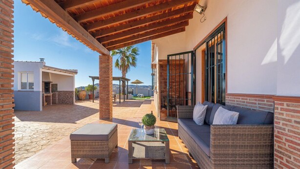 A covered outdoor patio with a wicker sofa, a glass table, and a brick wall. - Balcon de la Axarquia Almáchar by Ruralidays (Málaga)