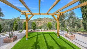 A pergola structure with a green lawn underneath, surrounded by trees and a distant hill.