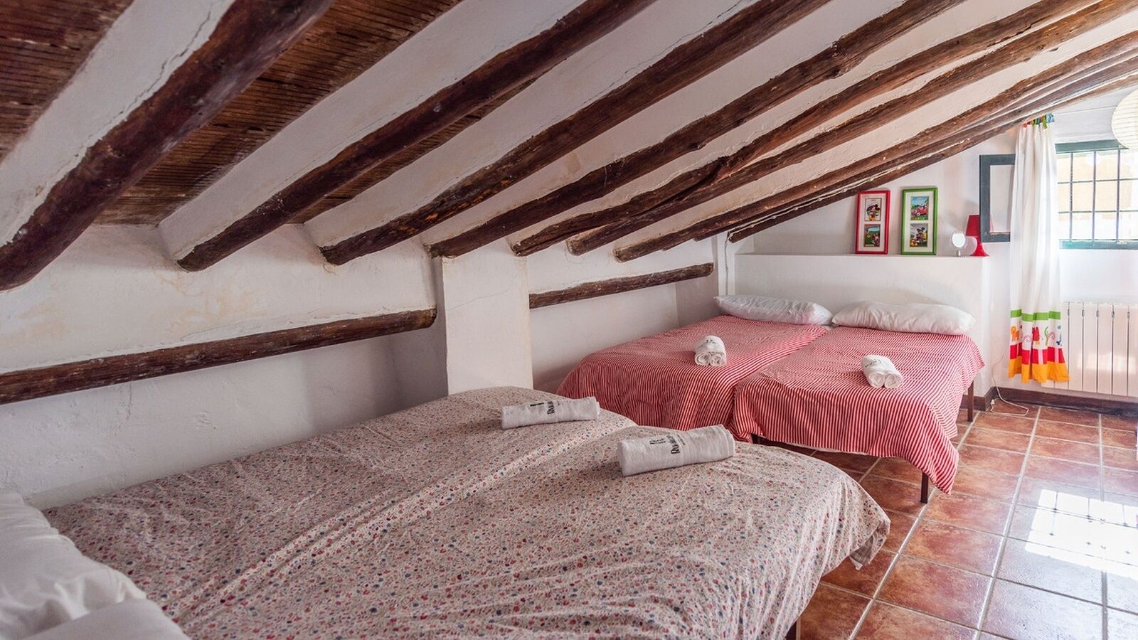 Une chambre avec deux lits, l’un recouvert d’une couvre-lit à motifs floraux et l’autre d’une couvre-lit à rayures rouges et blanches. Le plafond est orné de poutres en bois, et il y a une fenêtre avec un rideau.