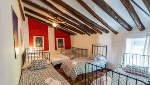 Ein Zimmer mit vier Betten, die in U-Form angeordnet sind, einer Holzbalkendecke und einem Fenster auf der rechten Seite.