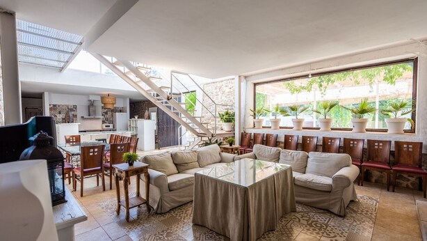 A spacious living area with a central coffee table, sofas, and a dining area with chairs. A staircase leads to an upper level, and potted plants are visible through the windows. - Cortijo La Vega Archidona by Ruralidays (Málaga)