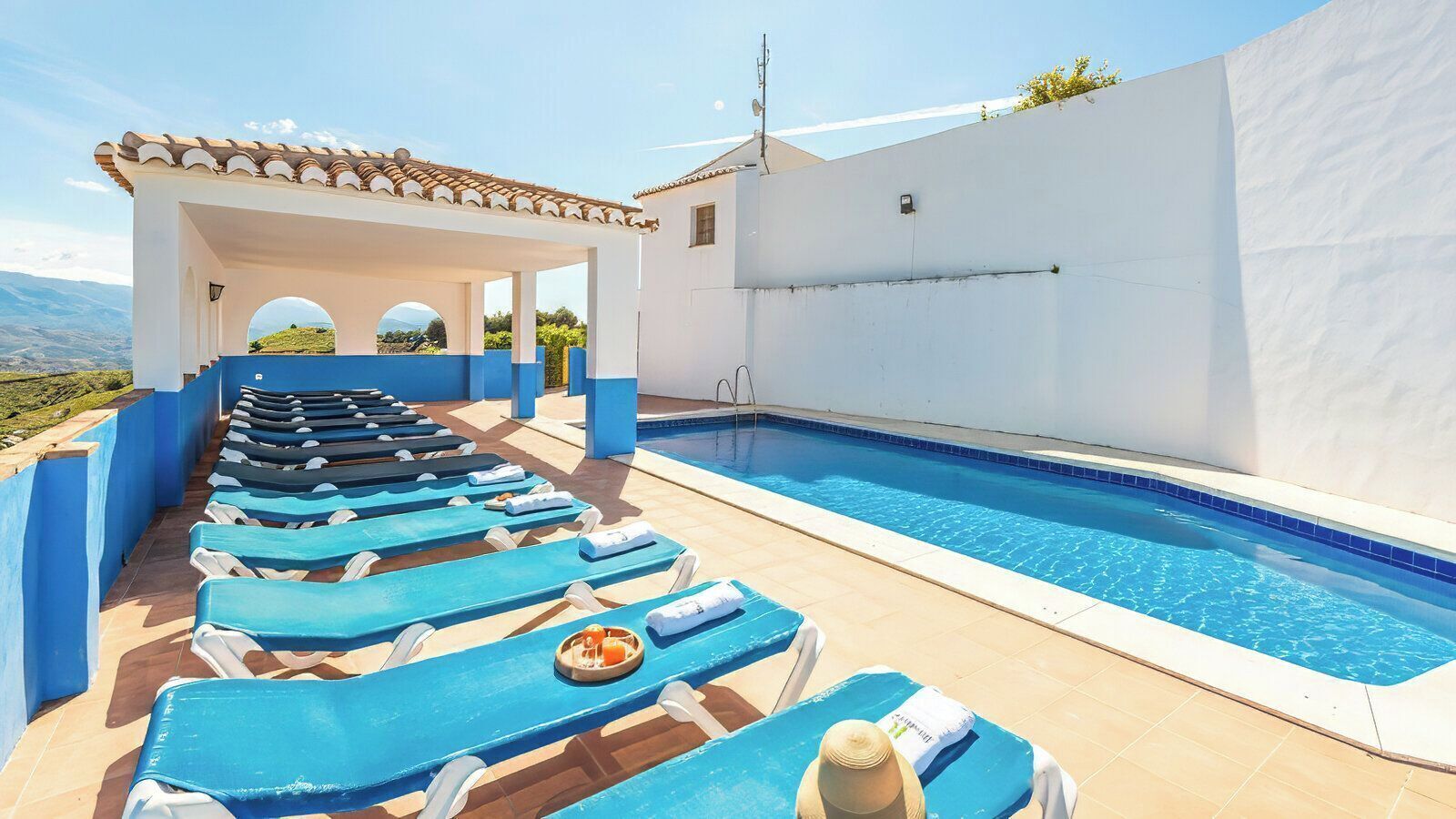 A pool area with blue lounge chairs and a white building with a tiled roof.